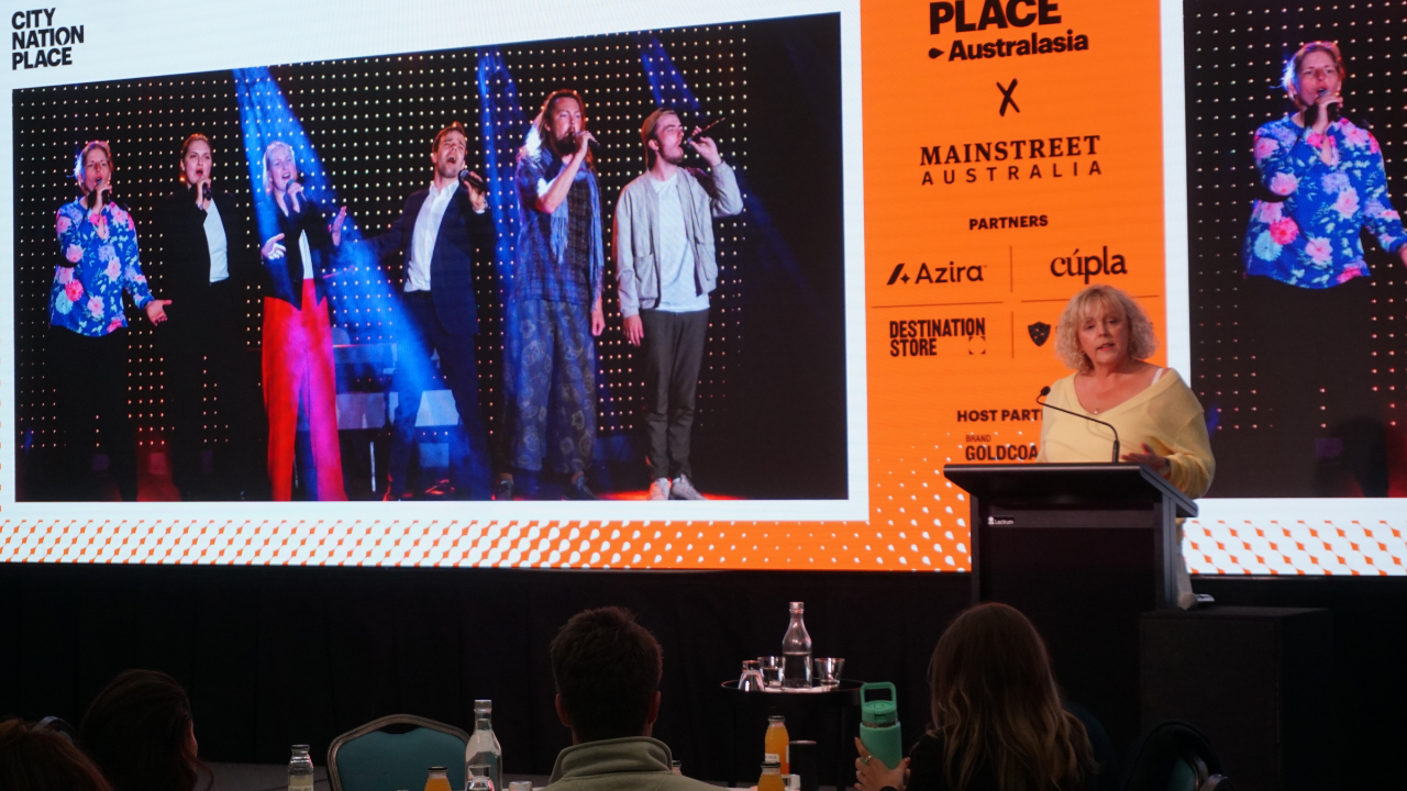 Clare Dewhirst presents at City Nation Place Australasia. She's standing at a lectern, and the screen shows an image of six actors with microphones from the City of Tartu's citizen engagement strategy.
