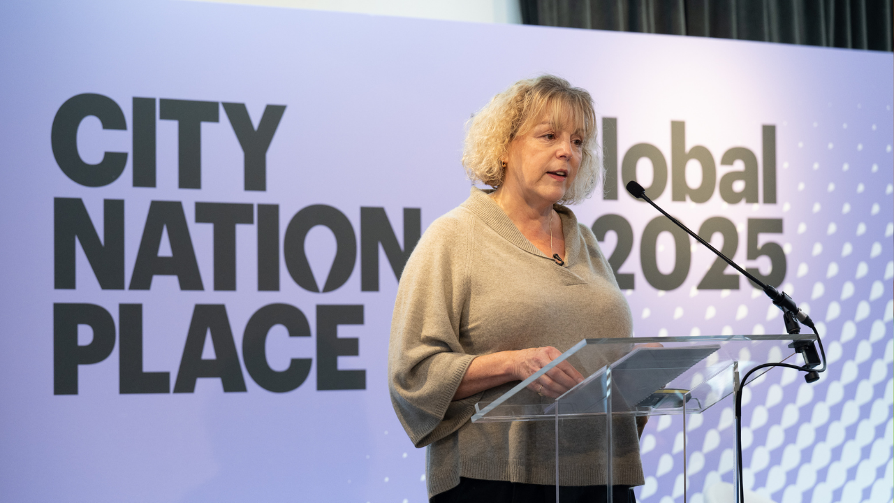 An image of Clare Dewhirst standing at a lectern in front of the purple backdrop for City Nation Place Global